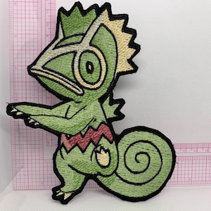 May include: Embroidered patch featuring a green chameleon-like creature with a yellow crest and red accents. The patch has a black border and is designed with detailed stitching. The creature is in a standing pose with its arms outstretched.
