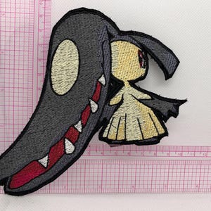 May include: Embroidered patch featuring a stylized character with a large, dark gray, toothy maw and a smaller, light-skinned figure with a yellow dress. The patch has black edging and is likely for clothing or accessories.