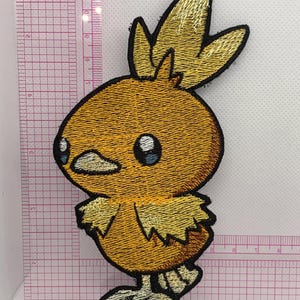 May include: An embroidered patch of a yellow chick-like creature with a three-pointed crest, large dark eyes, and light-colored feet. The patch is positioned against a clear plastic ruler showing a height of approximately 5.5 inches.