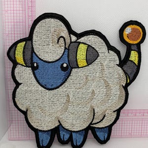 May include: An embroidered patch of a stylized sheep creature with cream-coloured wool, a blue face, and blue legs. It features black and yellow striped horns and a tail with a yellow orb at the end. A pink grid ruler shows the patch is 10 cm wide.