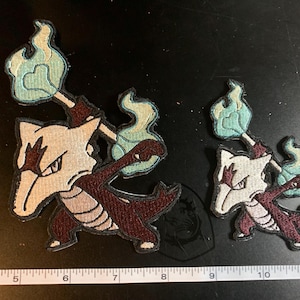 May include: Embroidered patches featuring a cartoon character with a bone-like head, dark red body, and a blue flame. The patches are in two sizes, with the larger one measuring approximately 4 inches in height.