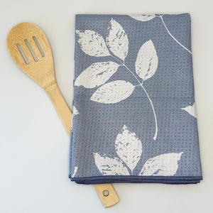 May include: A blue kitchen towel with a white leaf pattern. The towel is folded and a wooden spoon is lying on top of it.
