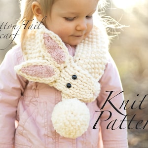 May include: A white and pink knitted bunny scarf with black button eyes and a white pom-pom tail. The scarf is worn by a child and the text "Cotton Tail Scarf Knit Pattern" is visible in the image.