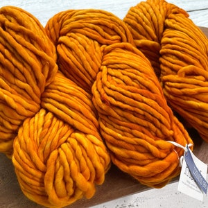 May include: Close-up of several skeins of vibrant orange merino wool yarn. The thick, chunky yarn is arranged in loose bundles, showcasing its texture. A tag is attached to one of the skeins. The yarn is ideal for knitting and other fiber crafts.