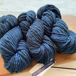 May include: Several skeins of thick, navy blue yarn with lighter blue highlights are arranged on a wooden surface. The yarn is chunky and appears soft. A tag with the text "150 AZUL PROFUNDO" is attached to one of the skeins.