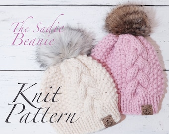 May include: Two cable-knit beanies with faux fur pom-poms. One is cream-colored, the other is light pink. The text "The Sadie Beanie Knit Pattern" is displayed in a script font. Each beanie has a small brown leather tag.
