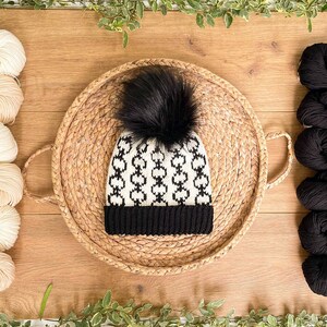 May include: A black and white patterned knit hat with a black pom pom on top. The hat is sitting on a woven straw tray. The tray is surrounded by white yarn balls and black yarn balls.