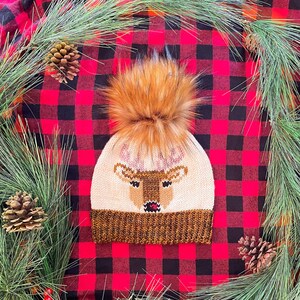May include: A knitted reindeer hat with a brown pom pom on a red and black buffalo plaid background.