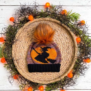 May include: A knitted witch hat with a brown, orange, and purple design. The hat has a black pom pom on top and a silhouette of a witch flying on a broomstick. The hat is sitting on a woven straw mat surrounded by orange and white lights.