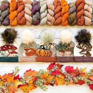 Puede incluir: Cinco gorros de punto con pompones en varios colores y patrones otoñales. Los gorros presentan diseños de una calabaza, un zorro, setas, bellotas y un ciervo.