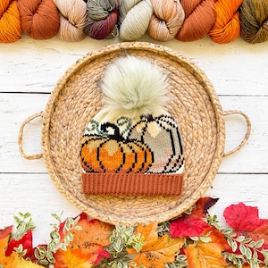 Könnte beinhalten: Eine gestrickte Mütze mit einem weißen Bommel und einem Muster aus Kürbissen und Blättern. Die Mütze ist braun, orange und weiß.