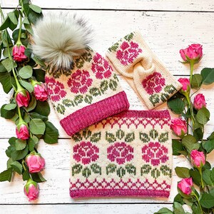 May include: A set of three knitted winter accessories featuring a floral pattern. The set includes a hat with a pom-pom, a headband, and a neck warmer. All three items are white with pink and green floral designs.