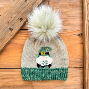 May include: A beige knit hat with a green and white gnome design and a white faux fur pom pom on top.