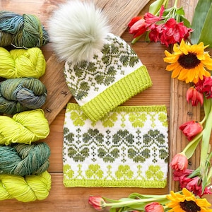 以下が含まれることがあります： 白と緑の花柄のニット帽とネックウォーマーのセット。帽子にはフェイクファーのポンポンが付いています。緑と黄色の糸が左側に並んでいます。セットは花が飾られた木製の表面に展示されています。