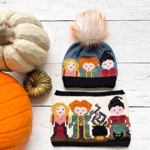 May include: A blue knitted hat with a white pom pom and a colorful embroidered image of the Sanderson sisters from the movie Hocus Pocus. The hat is paired with a matching white knitted scarf with a similar embroidered image of the Sanderson sisters.