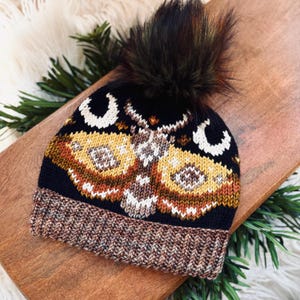 May include: A black knitted beanie with a brown and yellow moth design and a black faux fur pom pom.