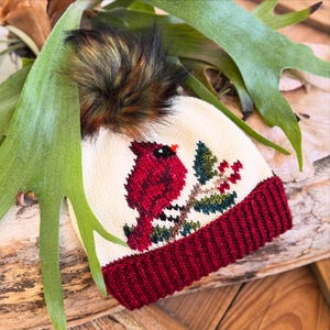 May include: Cream-colored knit hat with a red cardinal design and a burgundy brim. The hat features a brown and black faux fur pom-pom on top. The cardinal is perched on a branch with green leaves and red berries.