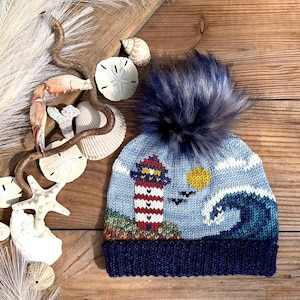 May include: A knit beanie featuring a lighthouse, a yellow sun, and a large blue ocean wave pattern. The hat has a dark blue ribbed cuff and a fluffy dark blue faux fur pom-pom on top. The design includes a red and white striped lighthouse.