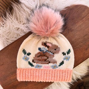 Può includere: Un cappello di lana con un pon pon rosa e un disegno di coniglio marrone. Il cappello è color crema con un bordo rosa e presenta fiori blu e foglie verdi.