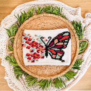 May include: A white knitted butterfly with red, pink, and green floral accents. The butterfly is on a woven straw-like round tray.