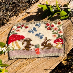 Puede incluir: Una diadema de punto de color crema con una escena de bosque que presenta dos conejos marrones, setas rojas y hojas verdes. La diadema tiene un borde morado claro y un borde superior gris.