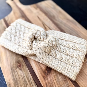 May include: Cream-colored knitted headband with a cable knit design. The headband is made of a soft, textured yarn and features a twisted knot detail at the center. The accessory is displayed on a wooden cutting board.