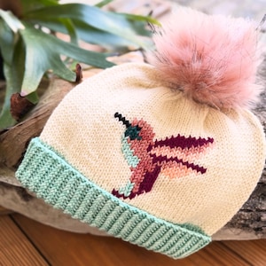 Puede incluir: Gorro de punto color crema con un diseño de colibrí en tonos turquesa, burdeos y melocotón. El gorro presenta un pompón de piel sintética rosa claro y un borde acanalado verde menta. Un accesorio de invierno.