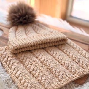Puede incluir: Un gorro de punto beige con un pompón marrón y una bufanda a juego. El gorro y la bufanda tienen un diseño de punto trenzado. El conjunto está hecho de un material suave y cálido, ideal para el invierno.
