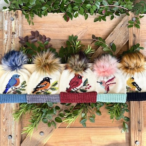 May include: Five cream-colored knit hats with colorful bird designs and faux fur pom-poms. Each hat features a different bird, including a bluebird, robin, cardinal, hummingbird, and goldfinch. The hats have different colored brims and are displayed on a wooden background.