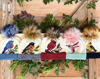 May include: Five cream-coloured knitted hats with colourful bird designs and faux fur pom-poms. Each hat features a different bird, including a bluebird, robin, cardinal, hummingbird, and goldfinch. The hats have different coloured brims and are displayed on a wooden background.