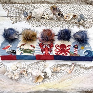 May include: Five knit beanies with pom-poms, each featuring nautical designs: a pelican, a crab, a lobster, an octopus, and a lighthouse with a wave. The hats are arranged in a row, showcasing detailed marine-themed patterns and textures.