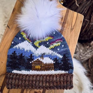 Puede incluir: Un gorro de invierno tejido con un pompón de piel sintética blanca. El gorro presenta un diseño de paisaje con montañas, árboles y una cabaña, en tonos azules, blancos, marrones y amarillos. El borde es de color marrón oscuro.