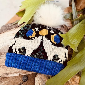 Puede incluir: Un gorro negro tejido con un diseño de lobo blanco, detalles azules y amarillos, y un pompón blanco esponjoso. El gorro tiene un borde acanalado azul y está sobre un fondo de madera natural y hojas verdes.