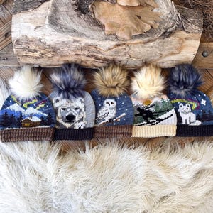 Op de afbeelding: Vijf gebreide wintermutsen met dieren- en landschapsontwerpen. Elke muts heeft een imitatiebont pompon en een bruine rand. De ontwerpen omvatten een ijsbeer, een uil en een landschap met het noorderlicht.