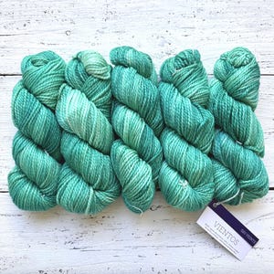 May include: Several skeins of teal-colored yarn are arranged on a white wooden surface. The yarn has varying shades of teal and green, creating a textured appearance. A small tag with the word "Vientos" is attached to one of the skeins.