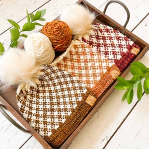 以下が含まれることがあります： 茶色、白、オレンジの糸で編まれた、格子模様のポンポン付きニット帽が2つ。帽子は木製のトレイの中にあり、背景には糸玉がある。