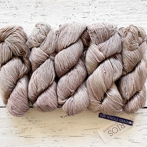 May include: A bundle of light brown yarn skeins, with a tag that reads "696 Whole Grain Rios". The yarn is arranged in a wavy pattern, with a soft, textured appearance. The background is a white wooden surface.