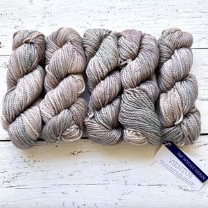 May include: Five skeins of gray and beige yarn are arranged in a row. The yarn has a textured appearance with subtle color variations. A tag in the lower right corner reads "696 Whole Grain" and "Vientos".