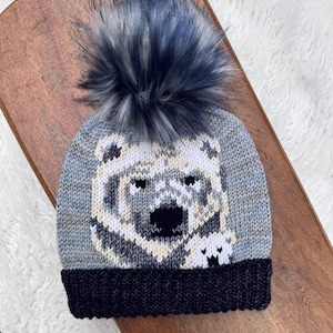 Puede incluir: Un gorro de invierno tejido con un diseño de oso polar. El gorro presenta un oso polar gris y blanco sobre un fondo azul claro, un borde negro y un gran pompón azul oscuro y esponjoso. Hecho de material suave y cálido.