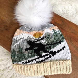 May include: A knitted winter hat with a white pom-pom. The hat features a mountain scene with a skier in black, a yellow sun, and green and white trees. The brim is a light beige color.