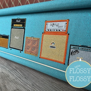 May include: A close-up of a cross-stitch embroidery of six guitar amplifiers on a blue fabric. The amplifiers are in different colors and styles, including a Marshall, a Fender, and a Vox. The embroidery is framed in a wooden hoop and has the text "sew FLOSSY FLOSSY" on the bottom right corner.