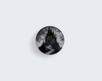 May include: A black and white button featuring a dark silhouette of a castle on a hill with a winding path leading up to it.