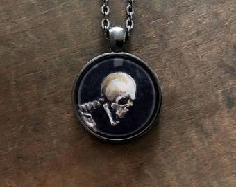 May include: A silver chain necklace with a round pendant featuring a black and white painting of a skull. The skull is painted in a realistic style with a white bone structure and black eye sockets.