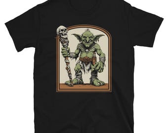 May include: Black t-shirt with a graphic of a green goblin-like creature holding a staff with a skull on top. The creature is wearing a brown loincloth and has a necklace with a skull pendant. The graphic is set in a brown frame.