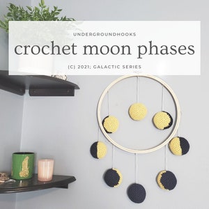 Peut inclure: Un cercle en bois avec des phases de lune en crochet jaune et noir suspendu à un mur. Le texte "crochet moon phases (c) 2021: Galactic Series" est au-dessus du cercle.