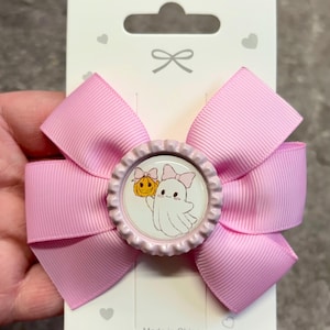 Könnte beinhalten: Eine hellrosa Haarschleife aus Ripsband mit einem mittigen Flaschenverschluss-Element. Das Element zeigt einen weißen Geist mit rosa Schleife, der einen kleinen orangefarbenen Kürbis mit rosa Schleife hält, befestigt auf einer weißen Karte.
