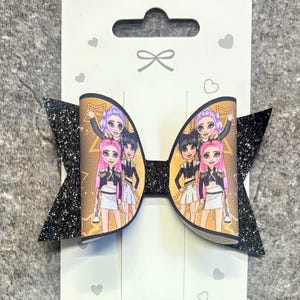 May include: A hair bow featuring a gold background with a repeating pattern of cartoon characters with pink, purple, and black hair, accented by black glittery side panels. The bow is attached to a white display card with the text Made in China.