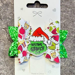 Könnte beinhalten: Eine Haarschleife mit einer weißen Kunstlederschicht mit einem wiederkehrenden Muster aus Grinch-Figuren und Weihnachtsgeschenken, akzentuiert durch hellgrüne Glitzer-Seitenteile und ein zentrales Emblem mit dem schwarzen Text Resting Grinch Face.