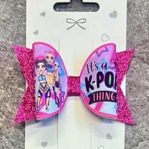 May include: A hair bow featuring a pink glitter finish on the outer edges and a central panel with a graphic print of three cartoon characters and the text It's a K-POP THING. The bow is attached to a white display card with small heart icons.