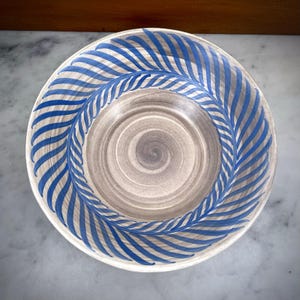 May include: A round ceramic bowl featuring a light grey centre with a subtle spiral pattern. The wide rim is decorated with a repeating blue painted feather or leaf motif that encircles the entire edge of the bowl, creating a distinct contrast.